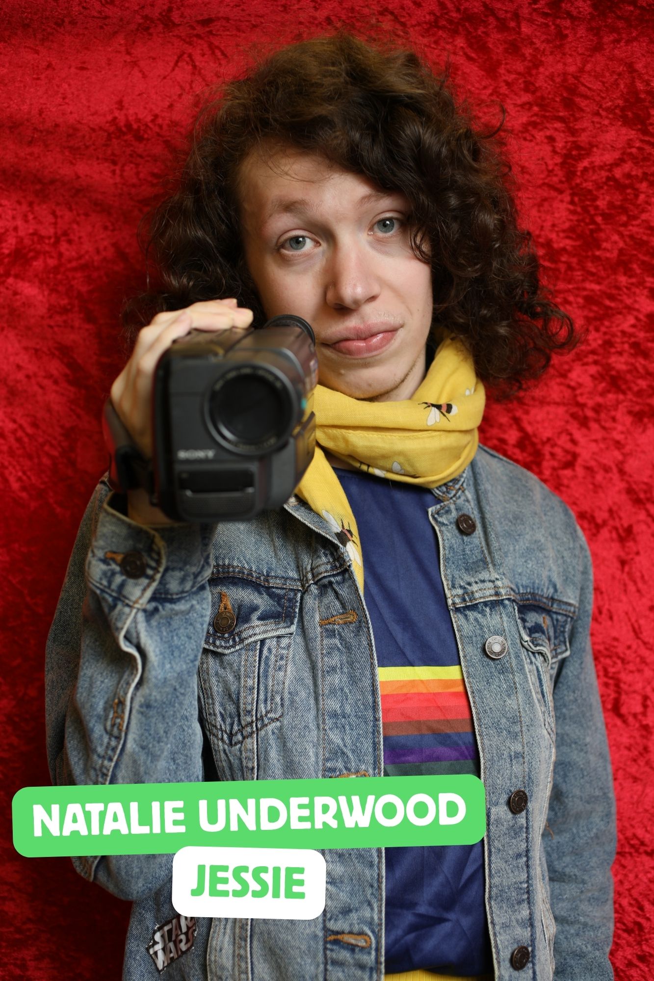 Nat, a white transwoman with brown, curly hair is playing Jessie. She is wearing a yellow skirt, a blue tshirt, and almost always holding a video camera.