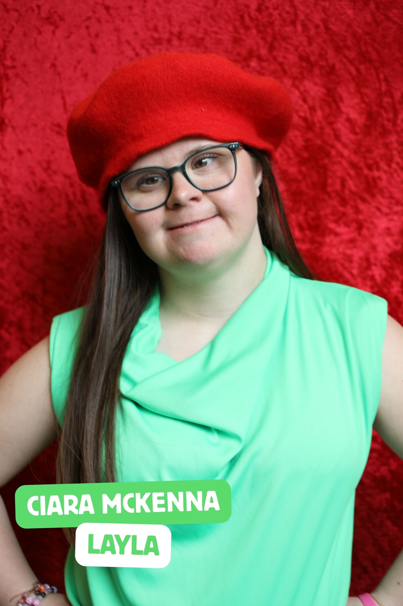 Ciara, a short, white woman who has Down’s Syndrome is playing Layla. She has long, brown hair and is wearing a bright green top, blue trousers, a red beret, and red-framed glasses..