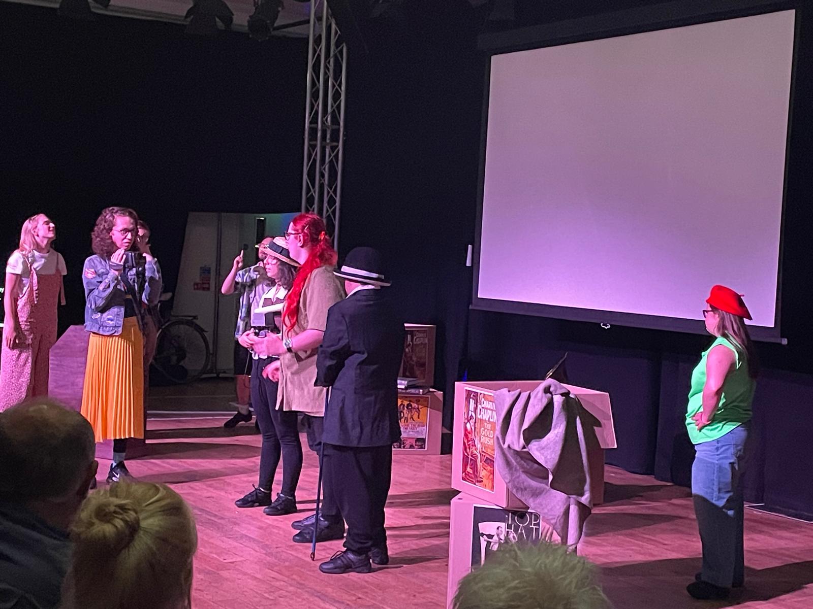 Members of the Reel to Real cast perform together on stage during the final Edinburgh show, with simple props and audience members visible in the foreground.