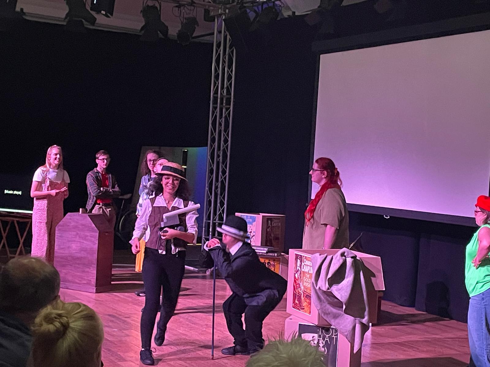 Performers act out a scene on stage during Reel to Real, including one performer in a dark suit and hat using a walking stick, with other cast members gathered around.