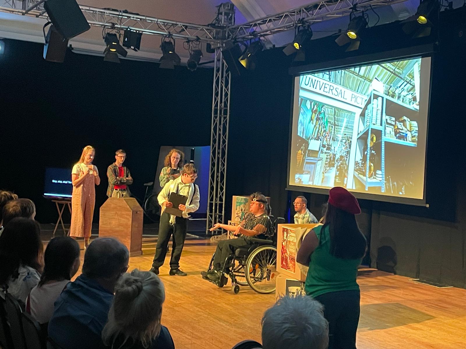 Several Reel to Real performers act on stage in front of the audience, with a large projected image of a film studio behind them and one performer using a wheelchair.