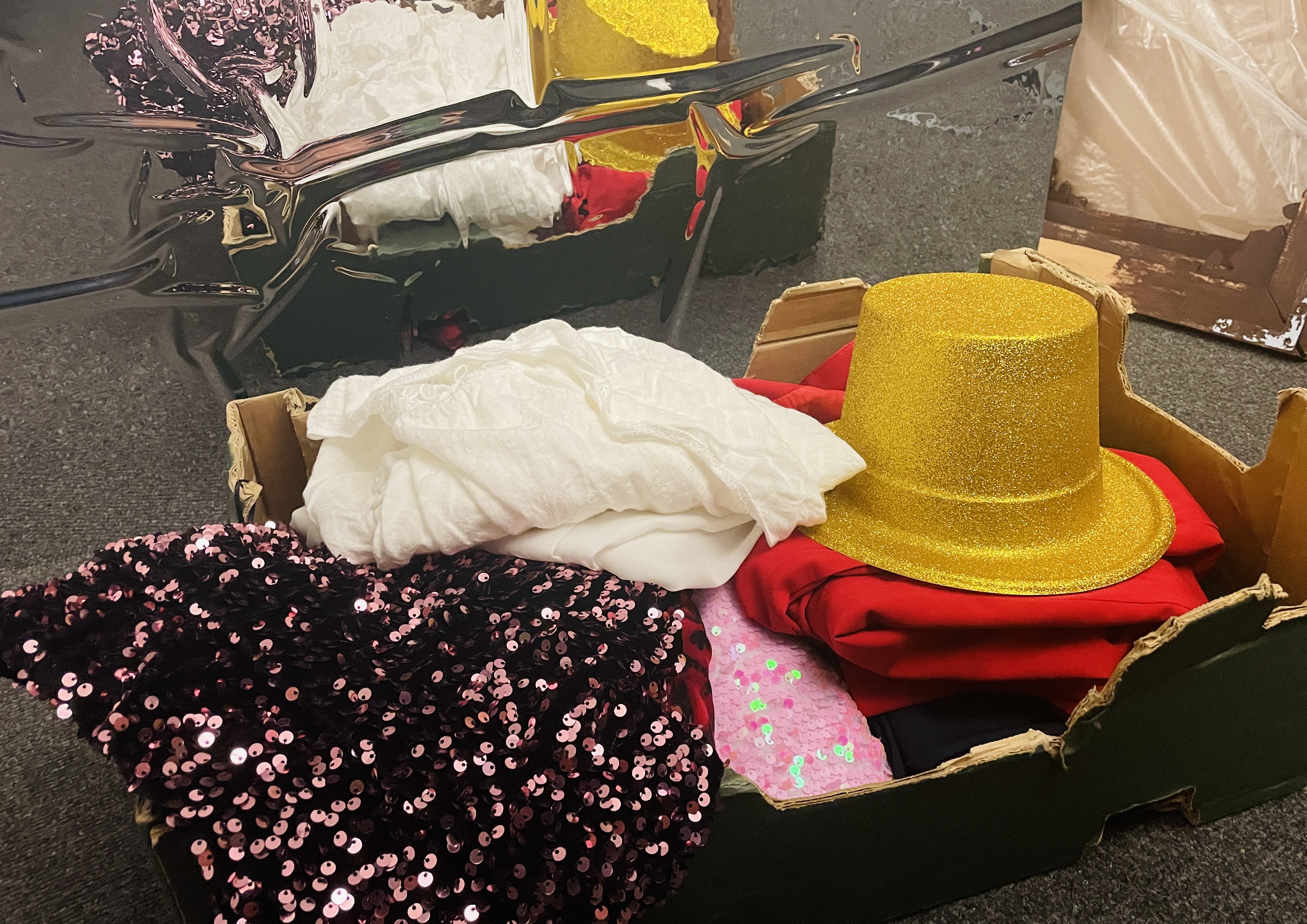 A box of costumes sit in front of a shiny surface. The box includes a pink sequin outfit and a gold, glitter top hat.