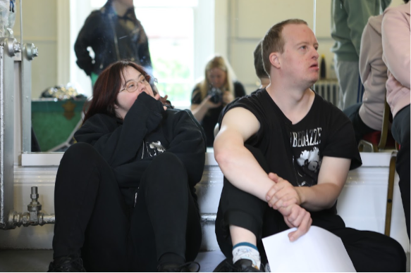 Ben, a white man with Downs syndrome, and another performer sit against a mirror watching a rehearsal intently. They look very focused. They are both wearing black t-shirt with the shiny Bedazzle logo.
