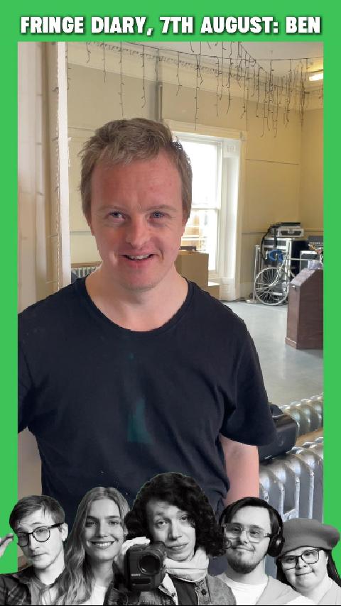 Ben, a white man in his 30's with Downs syndrome smiles at the camera. He is standing against a mirror in a rehearsal studio.