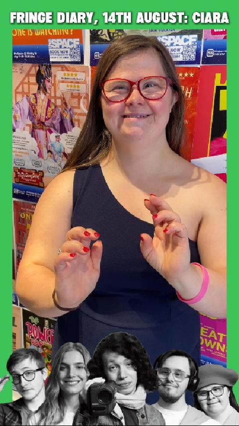 Ciara, a white woman with long brown hair, red-rimmed glasses and Down Syndrome is smiling at the camera. She is standing in front of a wall of theatre posters.