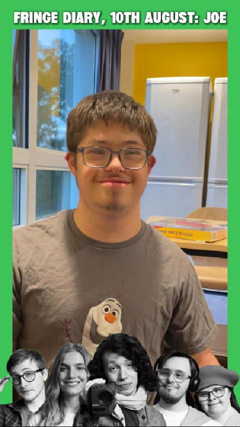 Joe, a white man with Down Syndrome, brown hair and blue-rimmed glasses, stands in the kitchen of an apartment and smiles.