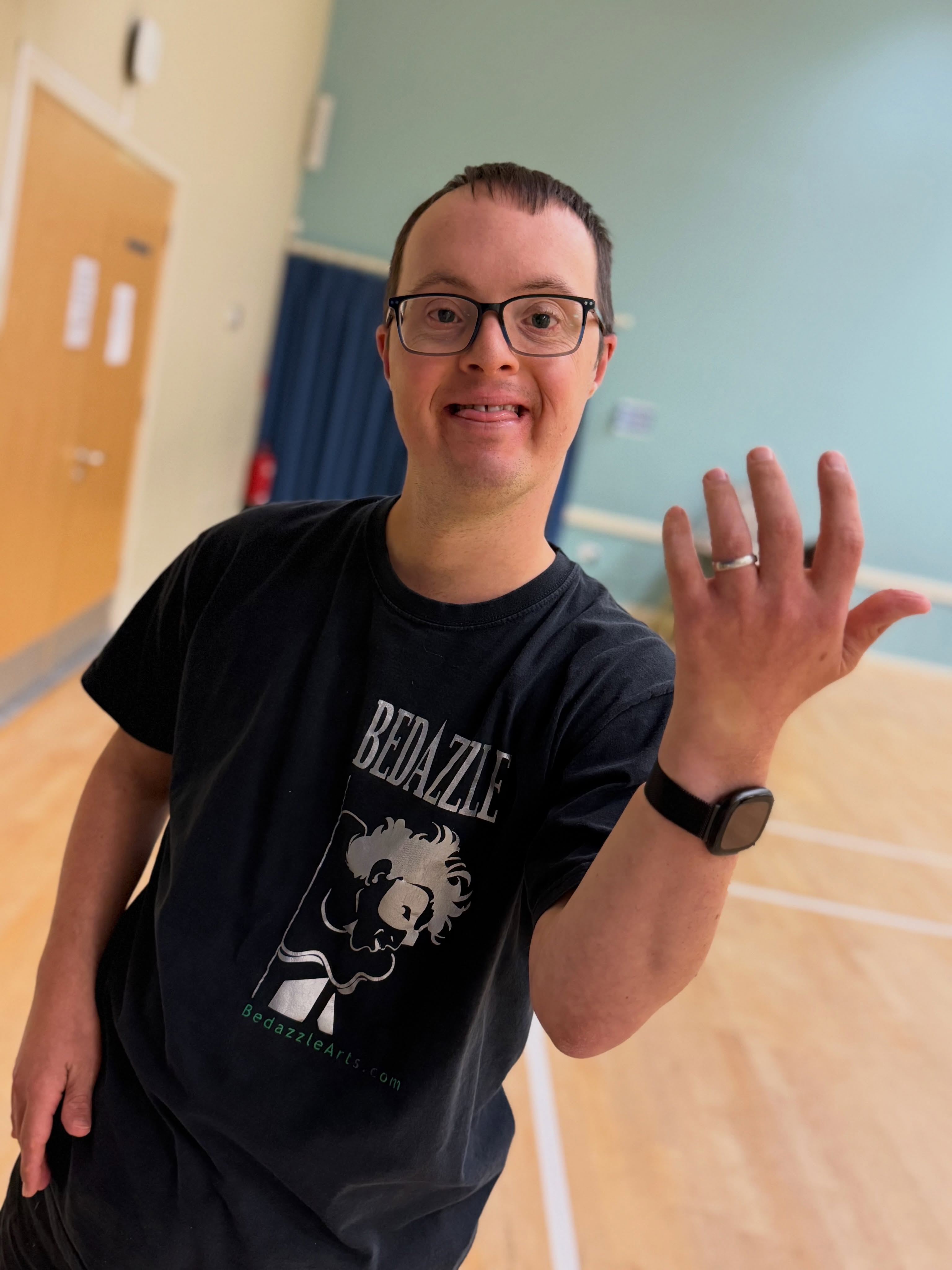 A smiling Bedazzle performer wearing a black Bedazzle T-shirt holds up his left hand to show his wedding ring during a community theatre session.