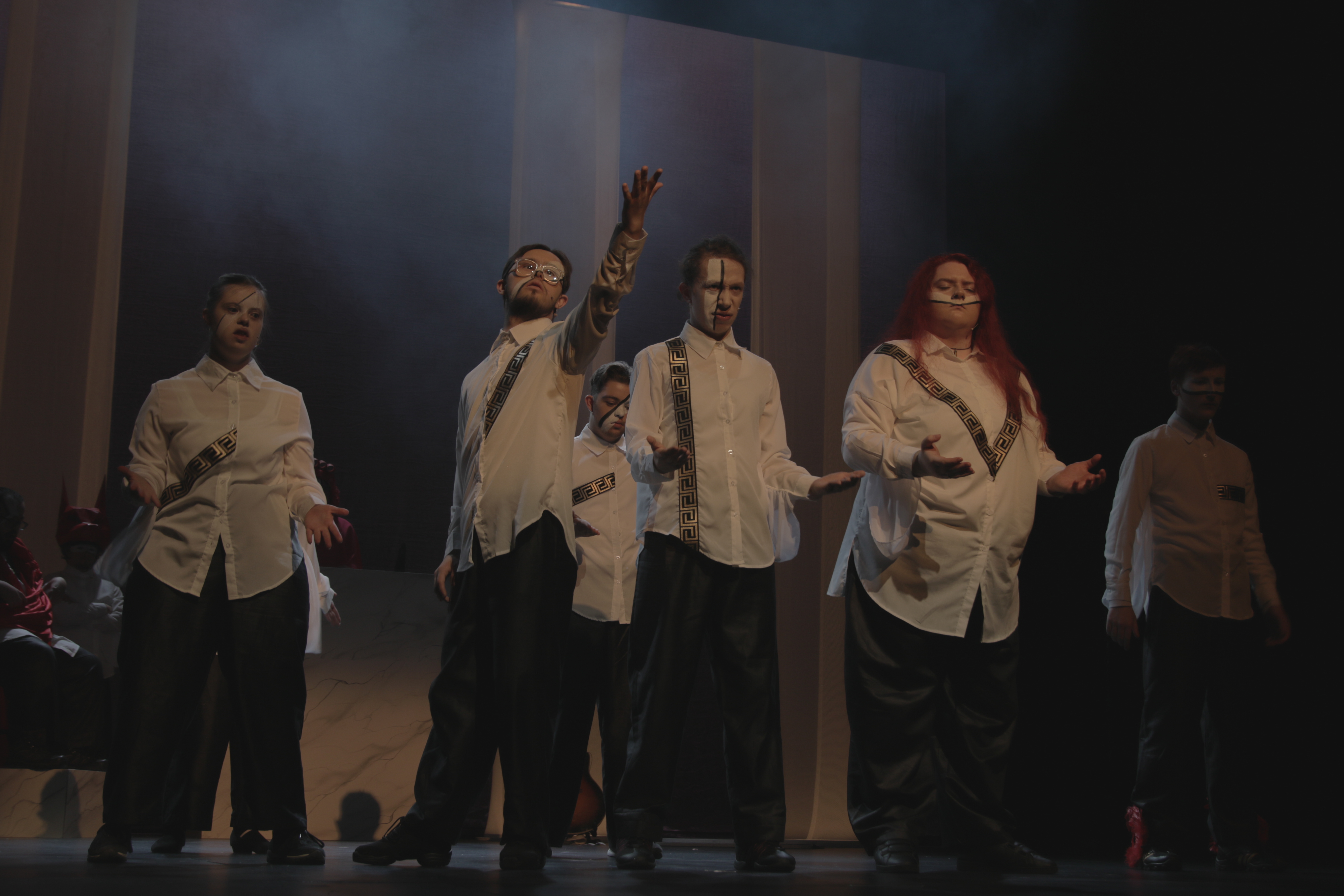 Stage photo from Mythic Trials showing performers in white shirts with black Greek-style trim and dark trousers, standing in dramatic poses under low theatrical lighting, with a misty set behind them.