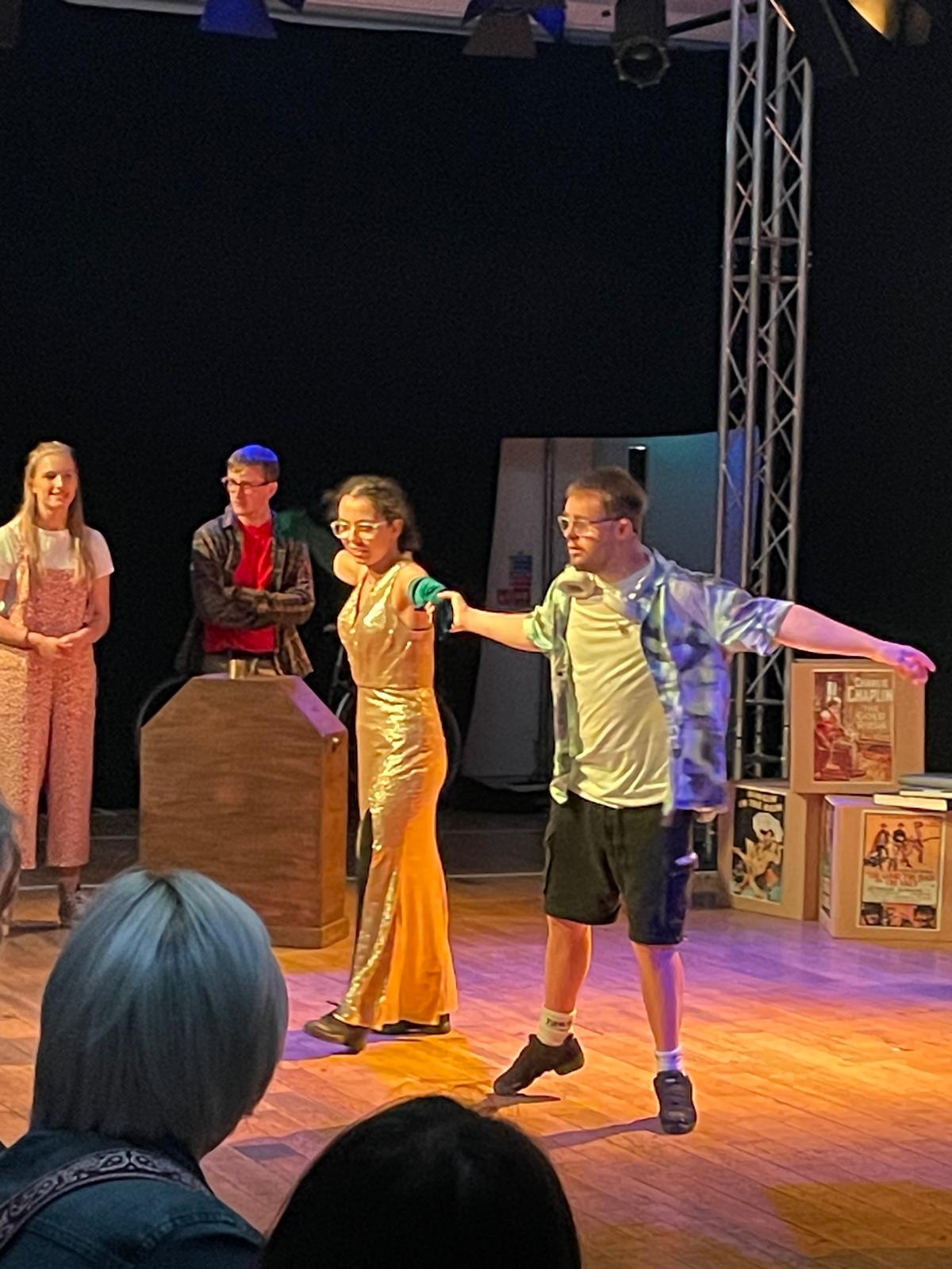 Two performers stand at the front of the stage during a scene, one wearing a gold costume and the other stretching an arm out dramatically, with other cast members behind them.