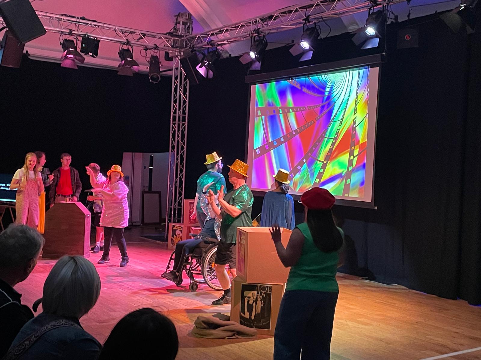 A group of performers in colourful costumes and hats perform together on stage, including a wheelchair user, with a brightly coloured film-themed image projected behind them.