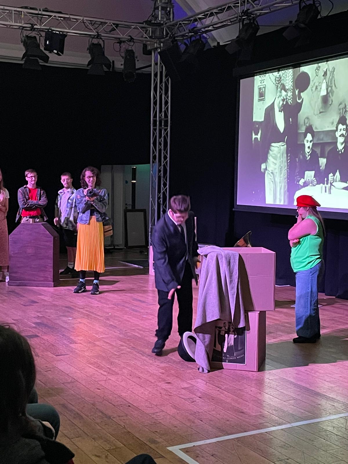 Performers act around simple stage props while a black-and-white film scene is projected behind them. Audience members can be seen watching from the side.