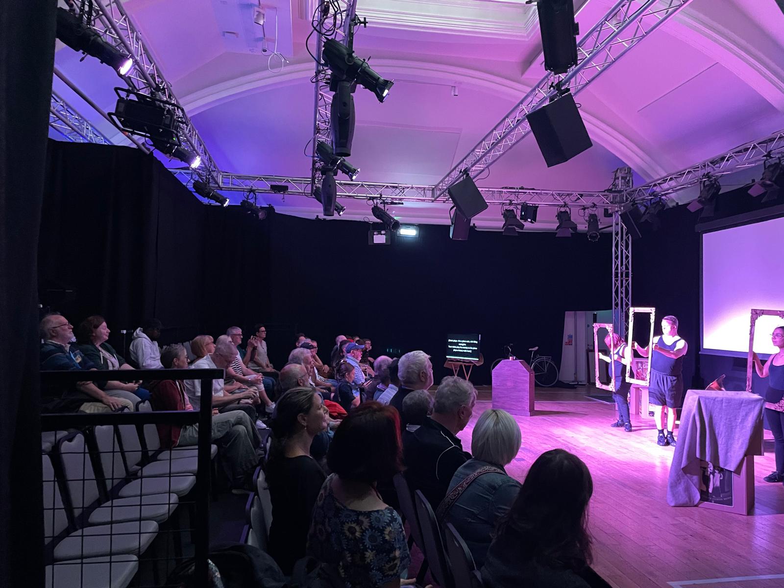 A wide view of the theatre shows a large seated audience watching performers on stage under purple lighting during Reel to Real.
