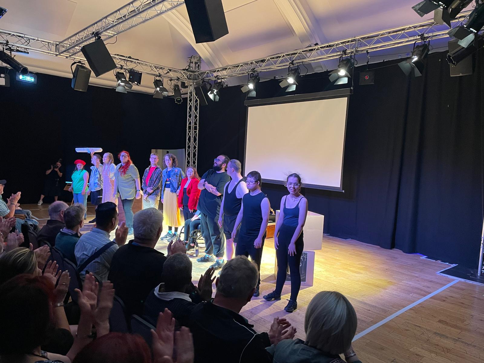 The Reel to Real cast stand together across the stage at the end of the performance while members of the audience applaud from their seats.