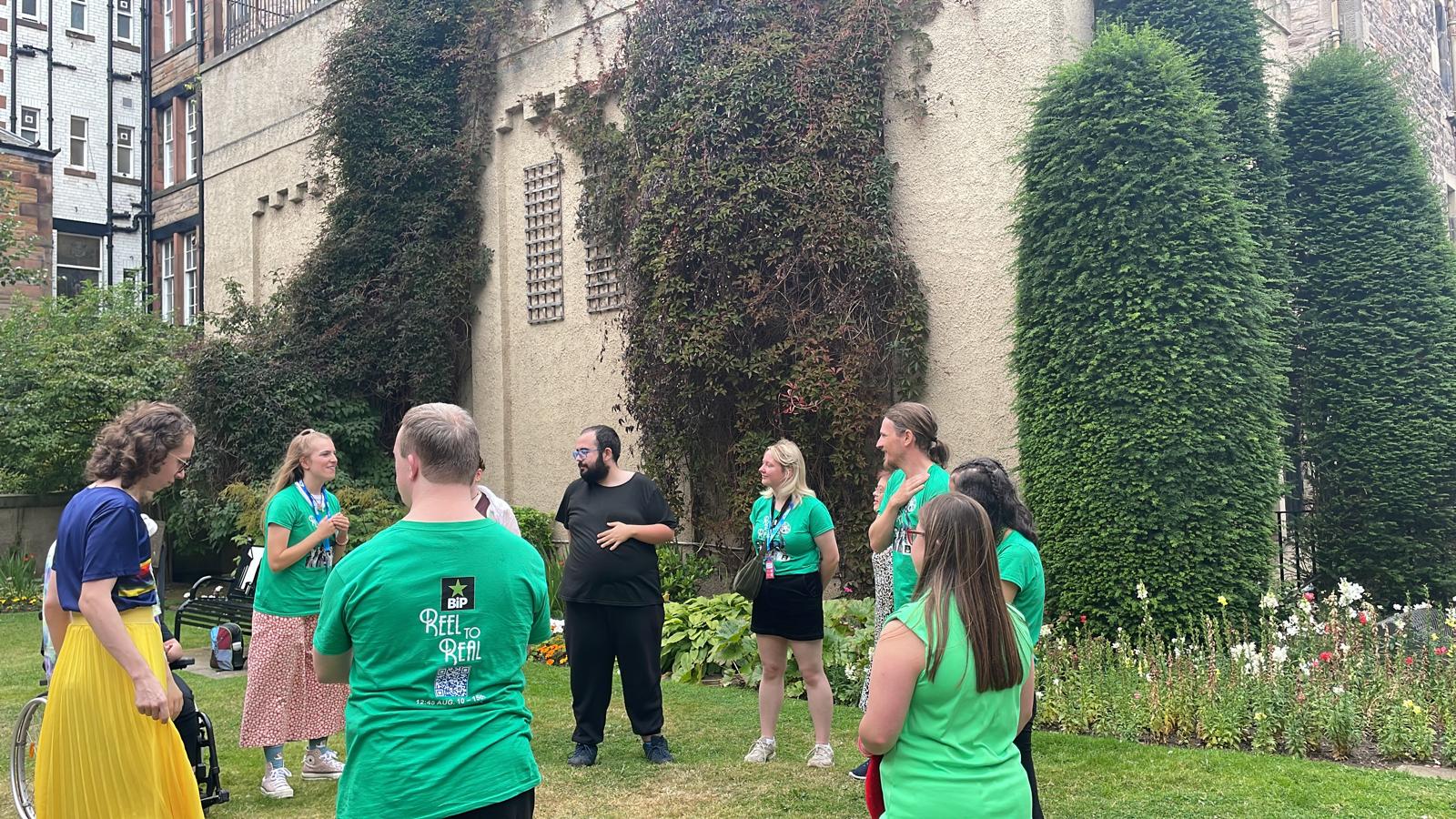 Members of the Reel to Real cast rehearsing together outside in a courtyard in Edinburgh. Several performers are wearing green show T-shirts, with others gathered around watching and taking part.