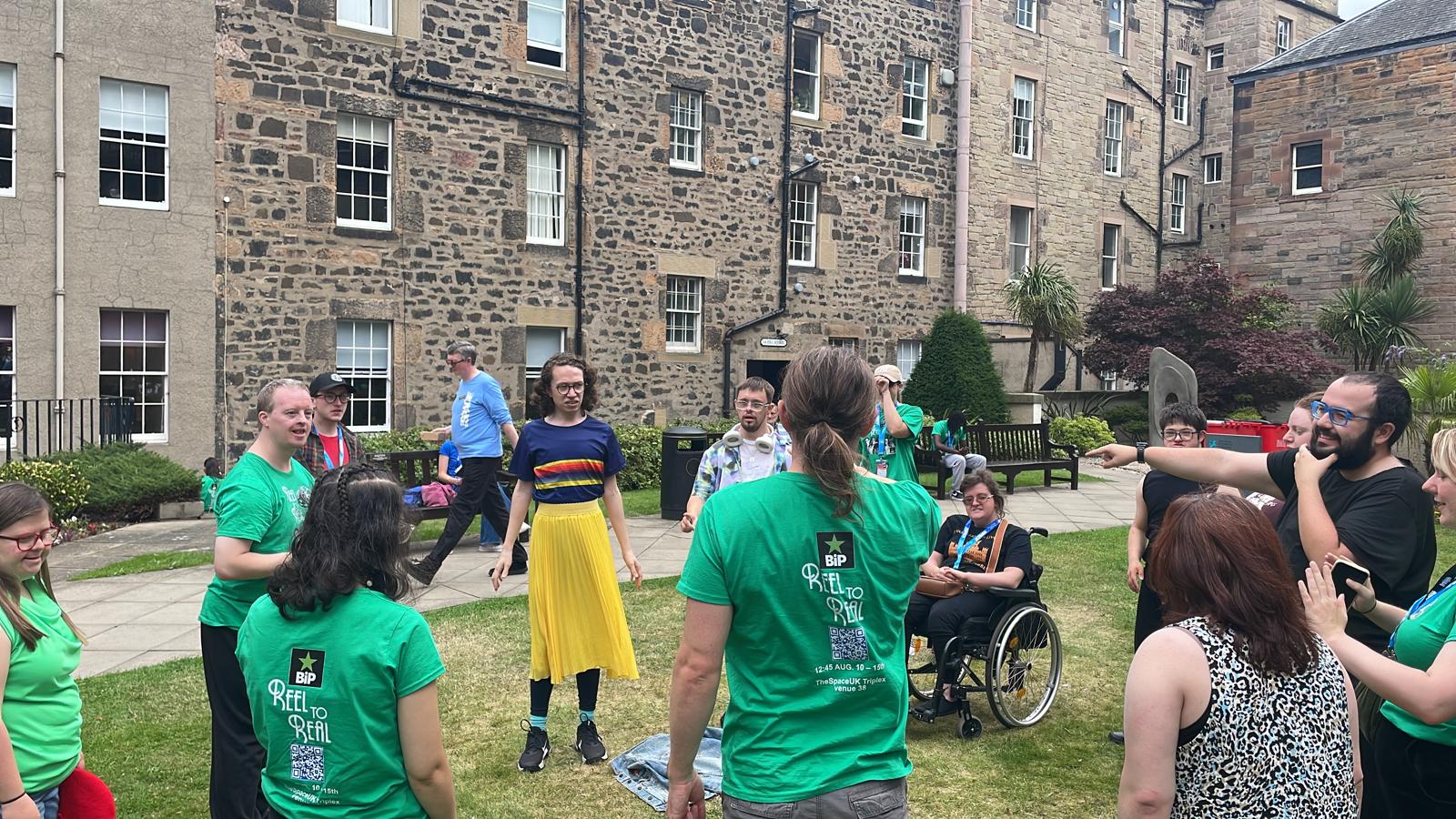 Members of the Reel to Real cast and team standing together during an outdoor rehearsal in a garden courtyard in Edinburgh, with several people wearing green show T-shirts.
