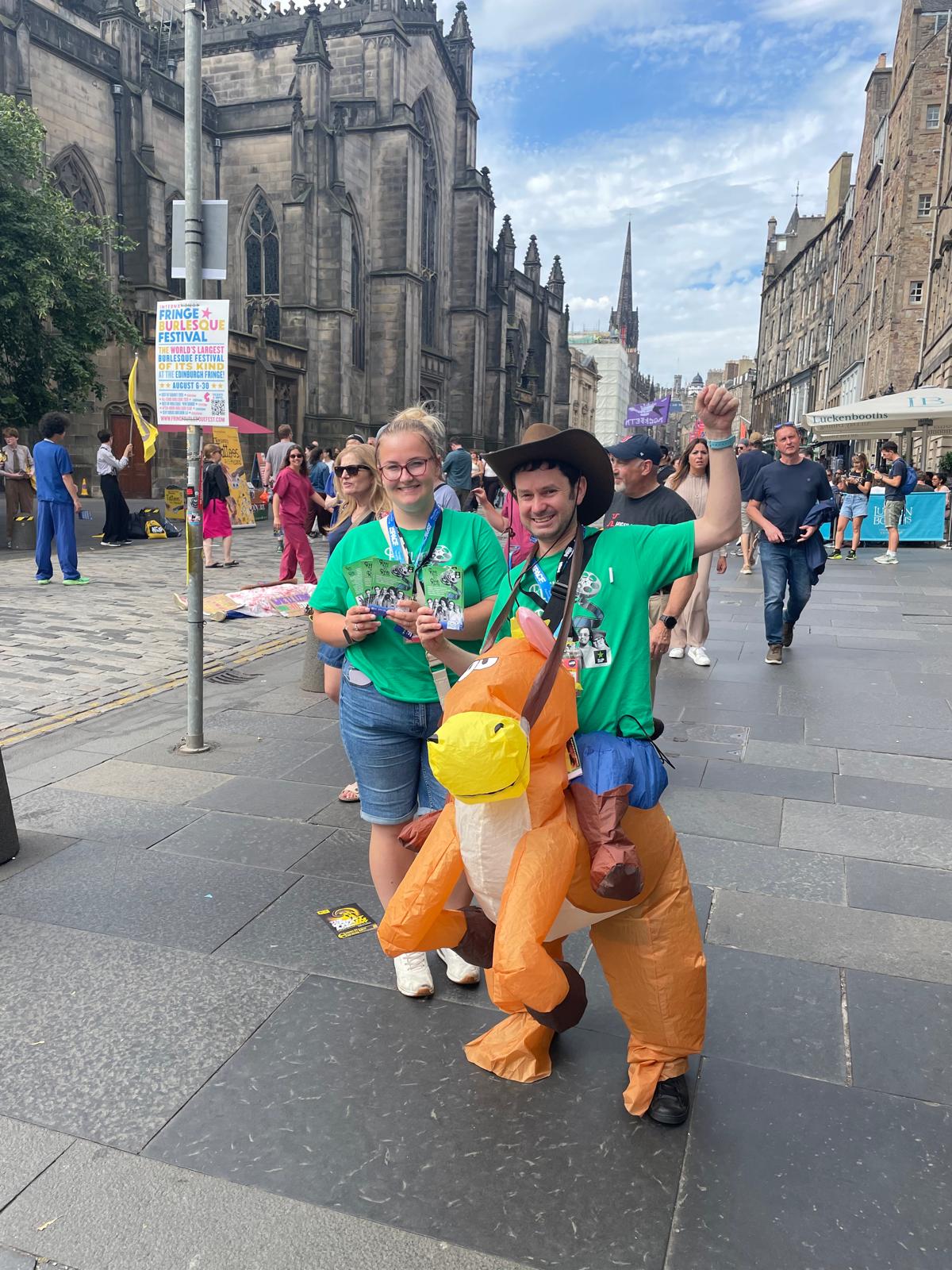 Courtney and Jacob handing out flyers for Reel to Real on a busy Edinburgh street during the Fringe. Both are wearing bright green show T-shirts, with Jacob dressed in an inflatable dinosaur costume and raising one arm in the air. Crowds and historic Edinburgh buildings can be seen behind them.
