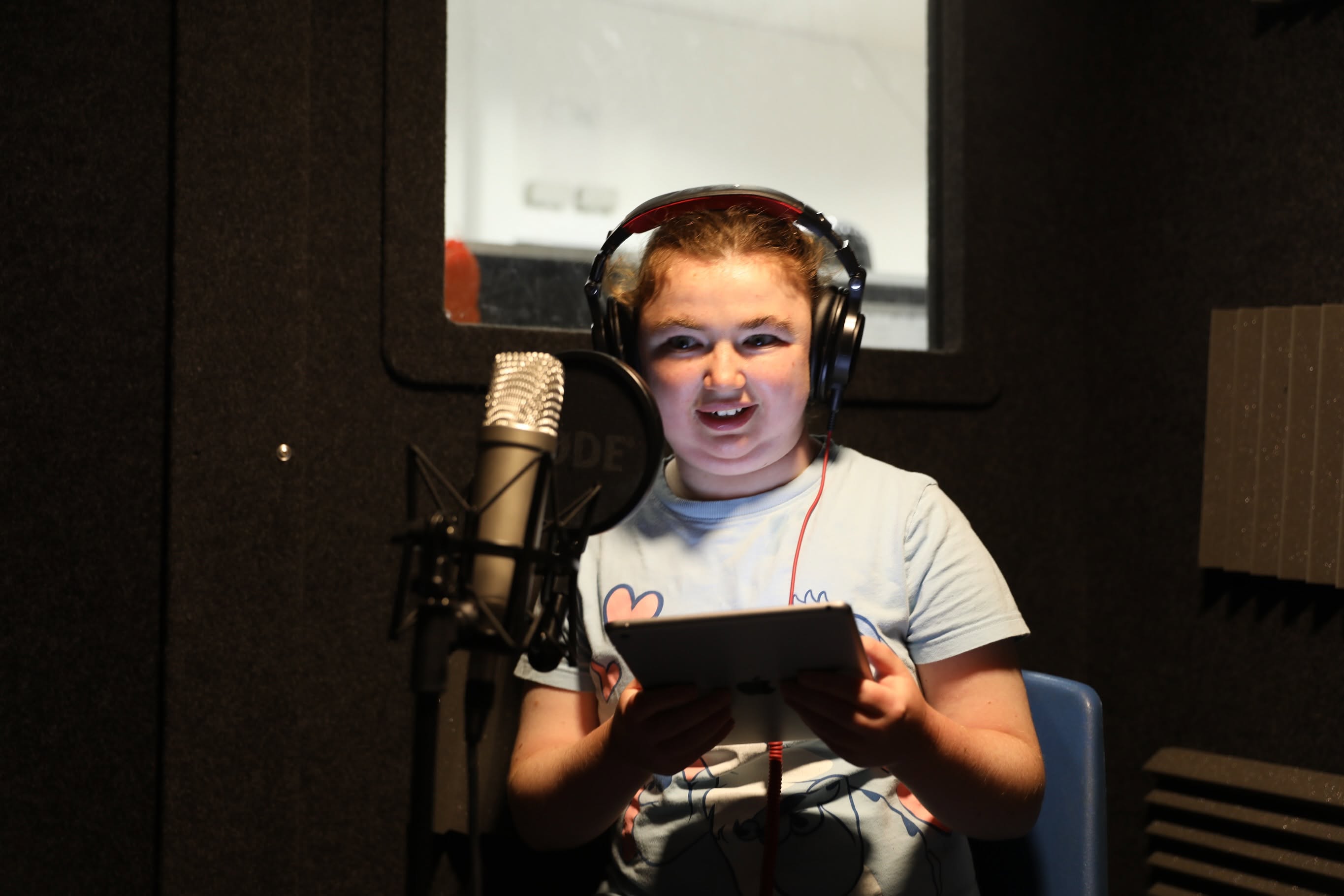 A Bedazzle performer wearing headphones smiles while reading from a tablet into a studio microphone.