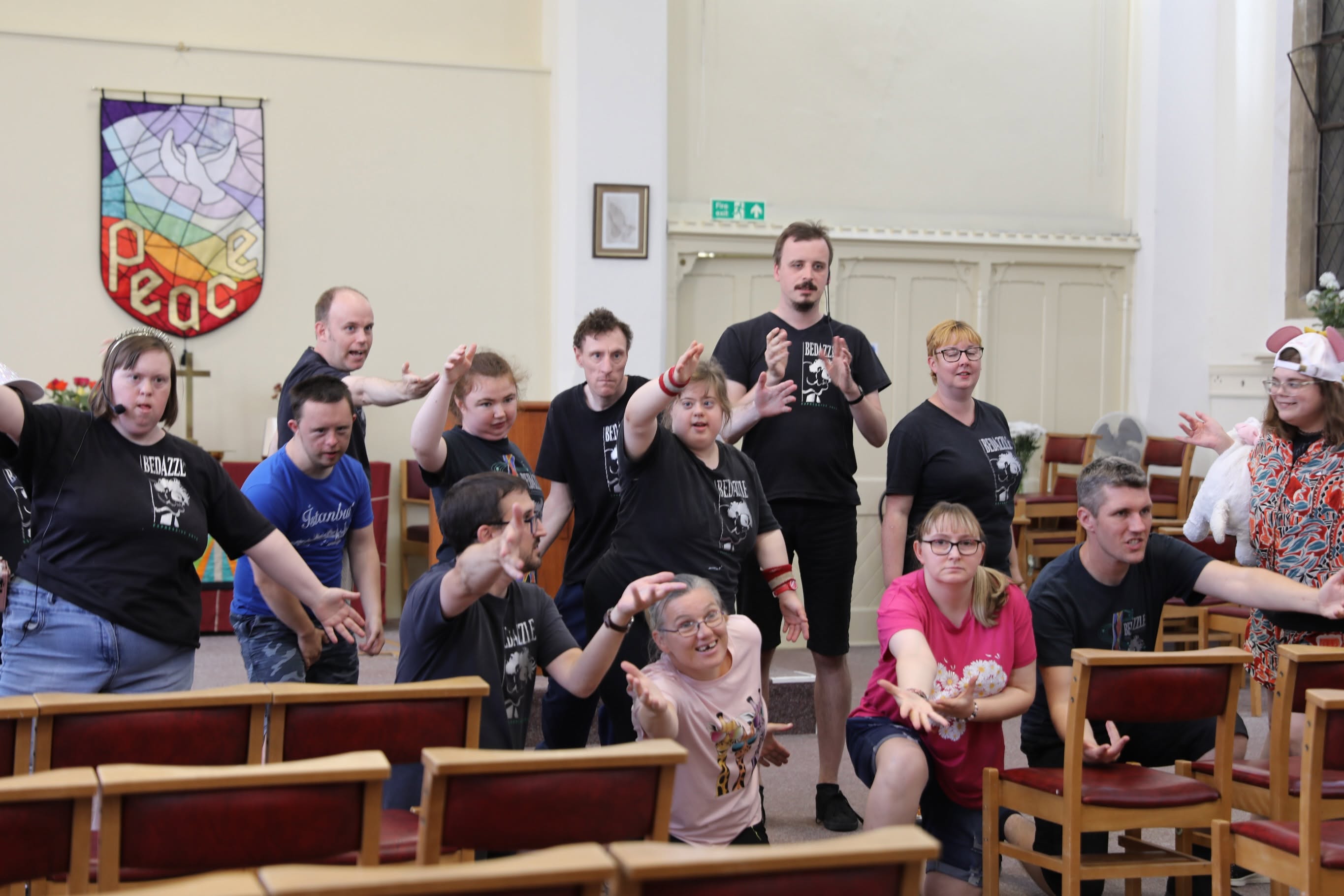 A group of Bedazzle performers rehearse together in a church hall, reaching and gesturing as part of a group performance.